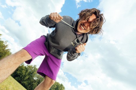 Woman with curly hair and wearing an orange T-shirt crouching on a rock with mountains in the&nbsp;background.