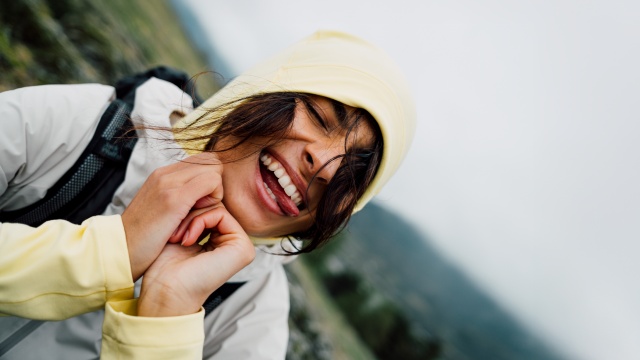 Close-up of a smiling woman in a yellow hood and light jacket, outdoors in a mountain landscape.
